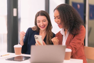 İki farklı iş kadını heyecan içinde ellerini kaldırıyor. Dijital tabletler ve ofislerdeki dizüstü bilgisayarlarda işbirliği hakkında sevinçle el kaldırıyorlar..