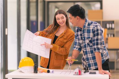 İki erkek ve kadın mimar planlar üzerine bir inşaat projesi hazırlıyorlar ve ofiste duran bir ev modeli emlak işiyle ilgili bir kız arkadaşı yerleştirmekten bahsediyorlar..