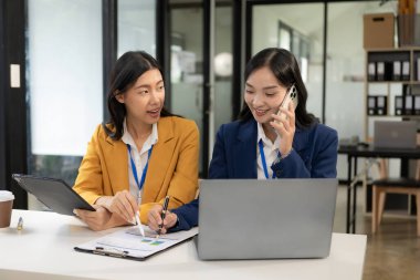 İki Asyalı kadın internette dizüstü bilgisayar ve akıllı telefonla çalışıyor. Ofisteki verimli bir toplantı sırasında iki bayan meslektaşın birlikte güldüğü neşeli bir an..