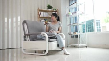 Asyalı kadın evdeki kanepede dinleniyor, sohbet uygulamasının tadını çıkarıyor, cep telefonunda metin yazıyor, canlı viral video izliyor, sosyal medya paylaşımlarını seviyor..