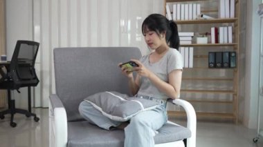 Asyalı kadın evdeki kanepede dinleniyor, sohbet uygulamasının tadını çıkarıyor, cep telefonunda metin yazıyor, canlı viral video izliyor, sosyal medya paylaşımlarını seviyor..