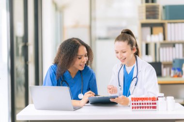 İki kadın bilimadamı, kan örnekleri ve test tüpleriyle bir laboratuvarda çalışıyorlar. Araştırmalarını sürdürürken, çözeltileri damlalardan gelen pipetlerle dolduruyorlar..