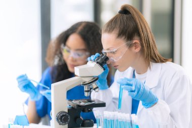 Koruyucu gözlüklü iki kadın tıp bilimcisi ve araştırma yaparken bir damlacıktan gelen bir pipetle çözelti dolu bir test tüpü..