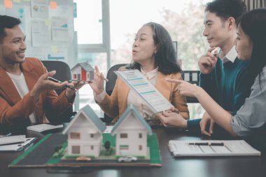 İş adamları, dizüstü bilgisayarlar ve mimari modellerle ofiste toplanıyorlar. Emlakçı, genç çifte ve iş kadınına ev modelini sunuyor.