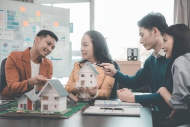 İş adamları, dizüstü bilgisayarlar ve mimari modellerle ofiste toplanıyorlar. Emlakçı, genç çifte ve iş kadınına ev modelini sunuyor.