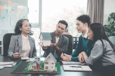 İş adamları, dizüstü bilgisayarlar ve mimari modellerle ofiste toplanıyorlar. Emlakçı, genç çifte ve iş kadınına ev modelini sunuyor.