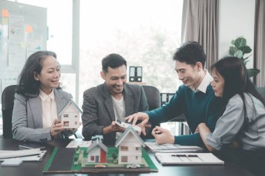 İş adamları, dizüstü bilgisayarlar ve mimari modellerle ofiste toplanıyorlar. Emlakçı, genç çifte ve iş kadınına ev modelini sunuyor.