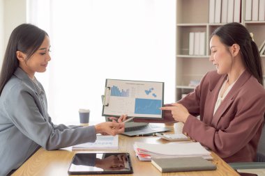 İki Asyalı kadın, mali grafik belgeleriyle çalışmak için hesap makinesi kullanarak kazanç ve iş gücünü artırmak için iş fikirlerini değiştirmek için konuşuyor..