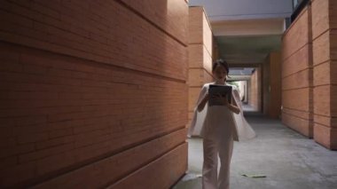Beyaz takım elbiseli bir tablet taşıyan Asyalı iş kadını kendinden emin ve günlük kıyafetlerle bağımsızdır. Gündüz vakti gülümseyen ve yürüyen - yavaş çekim