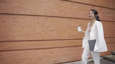 Beyaz takım elbiseli bir tablet taşıyan Asyalı iş kadını kendinden emin ve günlük kıyafetlerle bağımsızdır. Gündüz vakti gülümseyen ve yürüyen - yavaş çekim