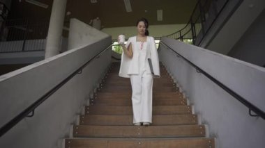 Beyaz takım elbiseli bir tablet taşıyan Asyalı iş kadını kendinden emin ve günlük kıyafetlerle bağımsızdır. Gündüz vakti gülümseyen ve yürüyen - yavaş çekim