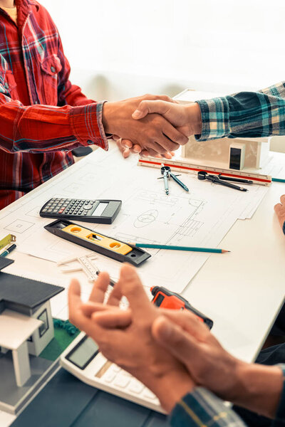 Architect and construction engineer holding hands, working as a team and cooperation concept after finishing agreement, close-up image with house plans on table
