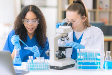 İki güzel kadın doktor, laboratuarda deney tüpleriyle bir masada oturuyorlar. Masada mikroskopla çeşitli sıvılarla dolu renkli cam şişeler.