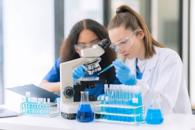 İki güzel kadın doktor, laboratuarda deney tüpleriyle bir masada oturuyorlar. Masada mikroskopla çeşitli sıvılarla dolu renkli cam şişeler.