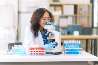 Güzel Afrikalı kadın doktor laboratuvar masasında test tüpleriyle oturuyor. Tahta bir masadaki çeşitli sıvılarla dolu renkli cam şişelerle.