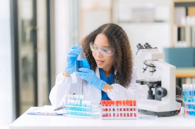 Güzel Afrikalı kadın doktor laboratuvar masasında test tüpleriyle oturuyor. Tahta bir masadaki çeşitli sıvılarla dolu renkli cam şişelerle.