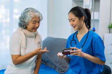 Üniformalı genç Asyalı doktor kıdemli kadın hastaya tıbbi tavsiyelerde bulunuyor ve hastane koğuşunda boncuklara oturuyor.