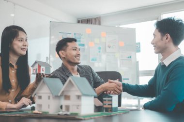 Emlakçı ve müvekkil, ahşap ev modeli ile imzalanan sözleşme üzerine el sıkışıyorlar. Belgeler, dizüstü bilgisayarlar ve insanlar konut projesi için müstakil bir ev inşa etmeyi tartışıyorlar.