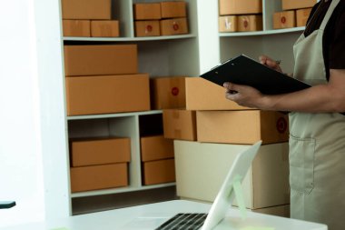 Genç adam bir çevrimiçi mağaza uzmanı. Paketleri postayla göndermek ya da taşımak için bir ürün kutusuyla çevrili bir dizüstü bilgisayarla masada oturmak. Resmi kapat..