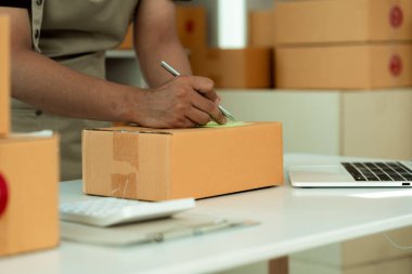 Genç adam bir çevrimiçi mağaza uzmanı. Paketleri postayla göndermek ya da taşımak için bir ürün kutusuyla çevrili bir dizüstü bilgisayarla masada oturmak. Resmi kapat..