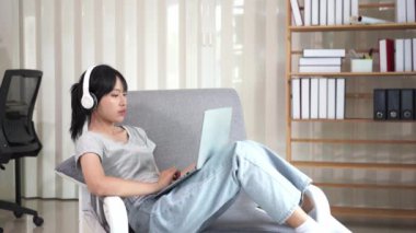 Asyalı bir kadın kanepeye oturmuş dizüstü bilgisayar kullanıyor, ekranda yazılmış metinlere bakıyor, bilgisayarla sohbet ediyor, internette, sosyal medyada, online alışverişte..