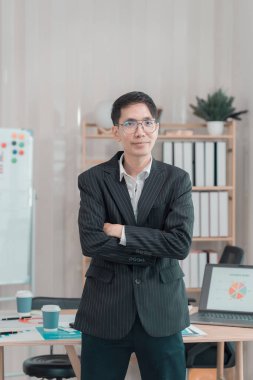 Ofiste başarılı bir iş adamı. Kollarını kavuşturmuş, gülümseyen ve bilgisayarla ofisin yanında duran kameraya bakıyor.