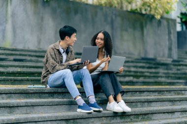 Çeşitli genç üniversite öğrencileri dışarıda dizüstü bilgisayar ve tabletlerle çalışarak teknoloji öğrenme kavramlarını tartışıyor..