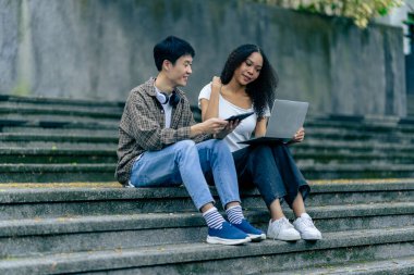 Çeşitli genç üniversite öğrencileri dışarıda dizüstü bilgisayar ve tabletlerle çalışarak teknoloji öğrenme kavramlarını tartışıyor..