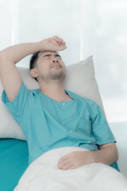 Asyalı endişeli bir erkek hasta. Eli alnında hastane yatağında yatıyor..