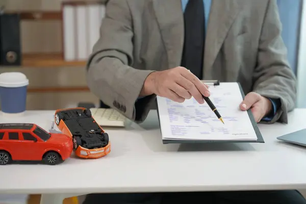 Araba sigortası ve finansal planlama konseptleri. Küçük oyuncak araba modeli. Araba sigortası belgelerinin yanına yerleştirilmiş. İşadamı kalem ve hesap makinesini ahşap masa üzerinde tutuyor.