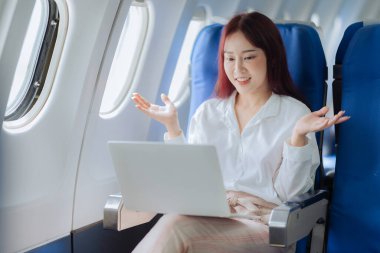 Mutlu Asyalı kadın uçakta oturuyor ve uçak penceresindeki uçuş sırasında dizüstü bilgisayar kullanıyor. Tatil yolculuğunun heyecanını ve heyecanını mükemmel bir şekilde aktarıyor..