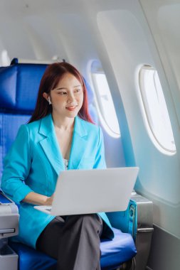 Mutlu Asyalı kadın uçakta oturuyor ve uçak penceresindeki uçuş sırasında dizüstü bilgisayar kullanıyor. Tatil yolculuğunun heyecanını ve heyecanını mükemmel bir şekilde aktarıyor..