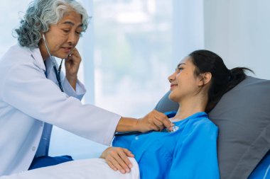 Yaşlı bir Asyalı kadın doktor hastane odasında genç bir hastayla birlikte bir panoya yazı yazıyor..