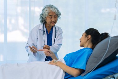 Yaşlı bir Asyalı kadın doktor hastane odasında genç bir hastayla birlikte bir panoya yazı yazıyor..
