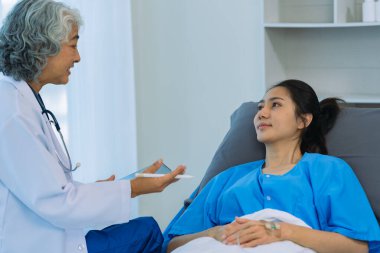 Yaşlı bir Asyalı kadın doktor hastane odasında genç bir hastayla birlikte bir panoya yazı yazıyor..