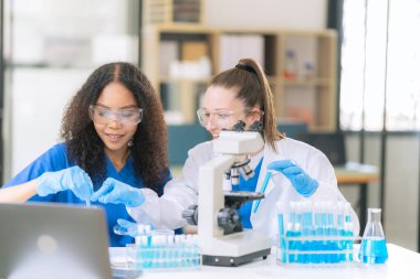 İki kadın bilim adamı modern bir laboratuvarda test tüplerindeki sıvıları inceliyor, özel koruyucu giysiler giyerken kimyasalları ve deneysel maddeleri inceliyor..