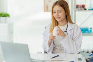 Laboratuvar önlüğü ve steteskopuyla gülümseyen Asyalı kadın doktor doktorun ofisinde online danışma masasının yanında oturuyor.