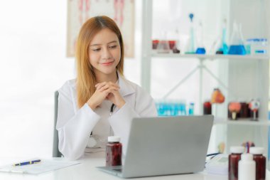 Laboratuvar önlüğü ve steteskopuyla gülümseyen Asyalı kadın doktor doktorun ofisinde online danışma masasının yanında oturuyor.