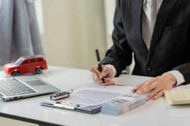 Küçük bir araba satıcısı hesap makineleri ve anahtarlarla çevrili bir masada otururken biri arka planda bir şeyler yazıyor. Araba satış konsepti.