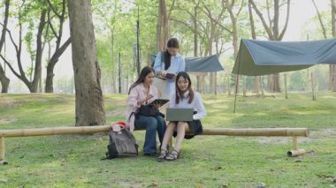 Bir grup Asyalı kız öğrenci akademik içeriği araştırmayı tartışıyor, tabletleri kullanıyor, parktaki bir ağacın altındaki not defterlerine not alıyorlar..