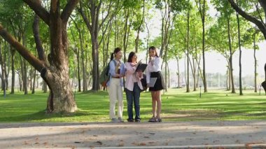 Bir grup Asyalı kız öğrenci akademik içeriği araştırmayı tartışıyor, tabletleri kullanıyor, parktaki bir ağacın altındaki not defterlerine not alıyorlar..