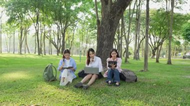 Bir grup Asyalı kız öğrenci akademik içeriği araştırmayı tartışıyor, tabletleri kullanıyor, parktaki bir ağacın altındaki not defterlerine not alıyorlar..