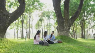 Bir grup Asyalı kız öğrenci akademik içeriği araştırmayı tartışıyor, tabletleri kullanıyor, parktaki bir ağacın altındaki not defterlerine not alıyorlar..