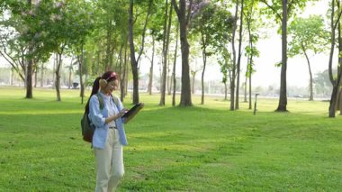Asyalı kız öğrenci akademik araştırmayı tablet kullanarak öğreniyor ve parktaki ağacın altındaki not defterine not alıyor..