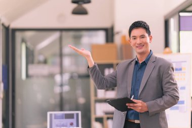 Asyalı işadamı dijital tablet tutuyor, çalışma alanında tek başına duruyor, kameraya bakıyor, modern web teknolojisini iş projesi yönetimi için kullanıyor.