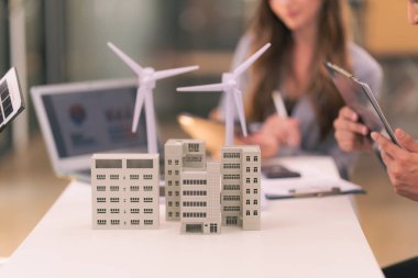 Rüzgâr ve güneş enerjisi kullanarak yenilenebilir enerji tasarlamak için güneş panelleri olan masaların üzerine ev modelleri ve planlarla rüzgar türbinleriyle buluşan mühendisler..