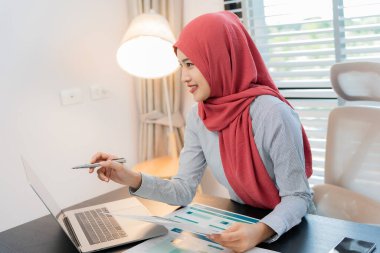 Ofiste dizüstü bilgisayarla çalışan tesettürlü Müslüman bir kadın, masadaki belgeler, planlama, analiz, mali rapor, iş planı, yatırım.