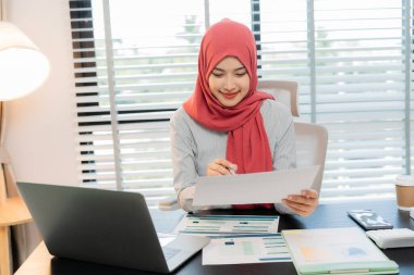 Ofiste dizüstü bilgisayarla çalışan tesettürlü Müslüman bir kadın, masadaki belgeler, planlama, analiz, mali rapor, iş planı, yatırım.
