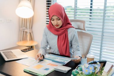Ofiste dizüstü bilgisayarla çalışan tesettürlü Müslüman bir kadın, masadaki belgeler, planlama, analiz, mali rapor, iş planı, yatırım.
