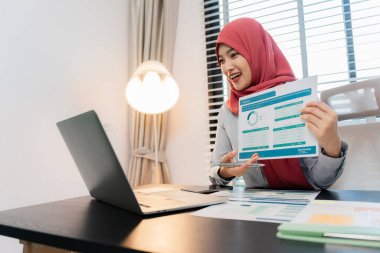 Ofiste dizüstü bilgisayarla çalışan tesettürlü Müslüman bir kadın, masadaki belgeler, planlama, analiz, mali rapor, iş planı, yatırım.